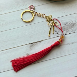 May include: A gold-toned metal keychain featuring a circular ring, a small pink flower charm, a decorative knot element, and a long red fabric tassel. The tassel is attached to the keychain with a red cord accented by small, translucent beads.
