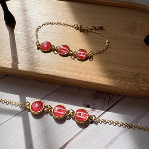 May include: Gold-colored chain bracelets with three red circular charms. Each charm has a white design. The bracelets are displayed on a wooden tray. The bracelets are delicate and elegant.
