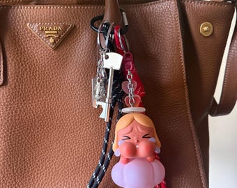 May include: A brown leather Prada handbag with a key chain featuring a crying cartoon girl, a key, and a bottle opener. The keychain has a red heart charm and a black and white lanyard.