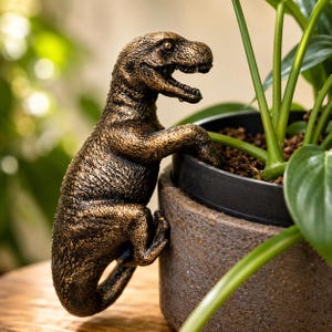 May include: A bronze-colored Tyrannosaurus Rex figurine with a textured, weathered finish. The dinosaur is depicted in a climbing pose, with its front claws hooked over the rim of a dark gray cylindrical plant pot containing a green plant.