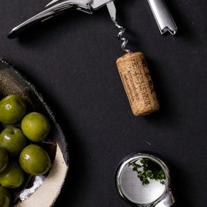 May include: A stainless steel corkscrew with a cork and a small dish of green olives. A small, round, silver-coloured magnifying glass is also in the image. The items are arranged on a black surface.