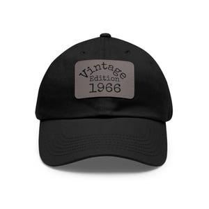 May include: Black baseball cap with a curved brim. A rectangular patch on the front displays the words "Vintage Edition 1966" in a serif font. The hat is made of a soft material and has an adjustable strap.