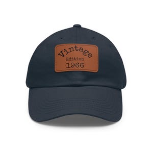 May include: A navy blue baseball cap featuring a rectangular brown leather patch centered on the front. The patch is embossed with the black text Vintage Edition 1966 in a typewriter-style font. The cap has a curved brim and stitched panels.