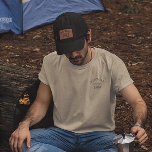 May include: A black baseball cap with a brown leather patch that reads "Vintage Edition 1968." The person is wearing a beige t-shirt with "Vintage Edition 1968" printed on the back. The person is sitting outdoors.