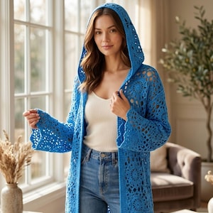 May include: A long, open-front, blue crochet cardigan with a hood. The cardigan features a floral pattern and bell sleeves. The person is wearing a white tank top and blue jeans. The background includes a window, a chair, and a plant.