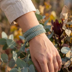 May include: A sage green macrame bracelet featuring a double-wrap design with a textured cord construction and a small knotted tassel detail at the end. The bracelet is crafted from woven thread and wraps twice around the wrist for a layered look.
