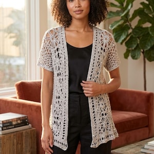 Puede incluir: Un cárdigan de crochet beige claro, de manga corta y abierto por delante. El cárdigan presenta un estampado floral y se lleva sobre una camiseta negra y pantalones negros. El cárdigan es un diseño ligero y aireado, perfecto para combinar.