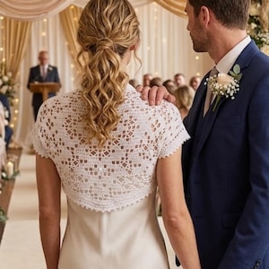 May include: A white crochet bolero with short sleeves and an intricate geometric pattern, worn over a smooth, sleeveless dress. The garment features a scalloped edge along the hem and neckline, providing a delicate, textured layer over the dress.