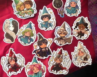 Puede incluir: Una colección de trece pegatinas de vinilo troqueladas con personajes estilo chibi con sombreros de mago, incluyendo varias figuras con cabello colorido, capas y efectos mágicos, además de una pegatina holográfica circular con un símbolo oscuro.