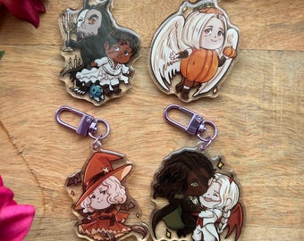 May include: Four clear acrylic keychains featuring illustrated chibi characters. Top left: a dark-haired figure with a candelabra and a child. Top right: a winged figure in a pumpkin suit. Bottom left: a witch in a hat. Bottom right: two figures embracing.