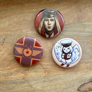 May include: Three circular pin-back buttons featuring distinct illustrations. The top button shows a person in a military-style cap and hood. The bottom left button displays a red apple with wings on a cross. The bottom right button depicts a winged figure.