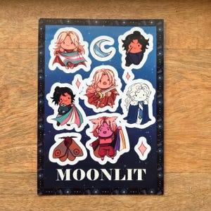 May include: A sticker sheet featuring several cartoon characters with various capes and accessories. The characters are set against a dark blue background with stars and the word "MOONLIT" at the bottom. The stickers have a white border.