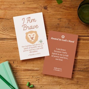 May include: Two cards with inspirational text and lion illustrations are on a wooden surface. One card reads "I Am Brave" with a lion graphic. The other card is brown and reads "Rooted in God's Word". A green notebook, pencil, and glass are also present.
