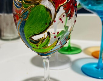May include: A hand-painted wine glass with a hummingbird design. The hummingbird is painted in shades of green, red, blue, and gold. The glass has a clear base and a red and orange design on the bottom.