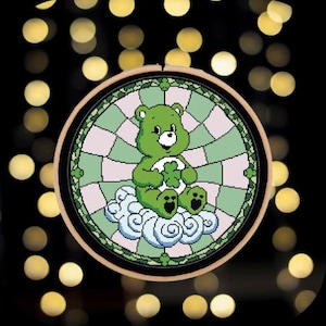 May include: A cross-stitch embroidery hoop featuring a green Care Bear holding a four-leaf clover, sitting on a cloud. The background is a stained-glass design with green and pink squares. The hoop is framed in a wooden ring.