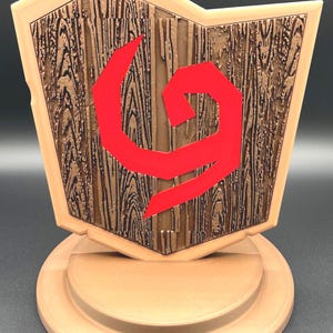 May include: A tan-colored decorative shield with a wood-grain texture and a red geometric design. The shield is mounted on a circular base. The shield has a light tan border.