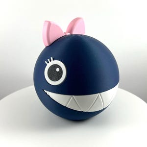 Könnte beinhalten: Eine marineblaue, kugelförmige Figur mit einer rosa Schleife oben. Die Figur hat ein weißes, zahnreiches Grinsen, ein weißes und schwarzes Auge und eine kleine Wimper. Das Objekt ist wahrscheinlich ein Dekorationsartikel oder ein Spielzeug.