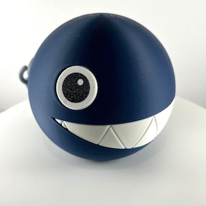 May include: A navy blue, spherical decorative item with a white, toothy grin and white eye details. The item has a textured surface and a small, gray loop attached to the top. The design is reminiscent of a cartoon character.