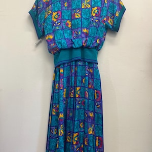 May include: A vintage teal dress with a geometric pattern in purple, yellow, and pink. The dress features short sleeves, a round neckline, and a gathered waist. The skirt is pleated, and the dress hangs on a wooden hanger.