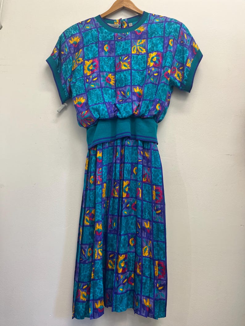 May include: A vintage teal dress with a geometric pattern in purple, yellow, and pink. The dress features short sleeves, a round neckline, and a gathered waist. The skirt is pleated, and the dress hangs on a wooden hanger.