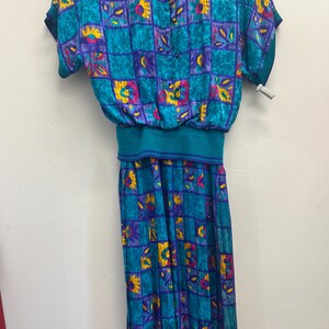 May include: A vintage teal dress with a square pattern in purple, yellow, and pink. The dress features short sleeves, a cinched waist, and a button-up back. The skirt is a long, flowing style.