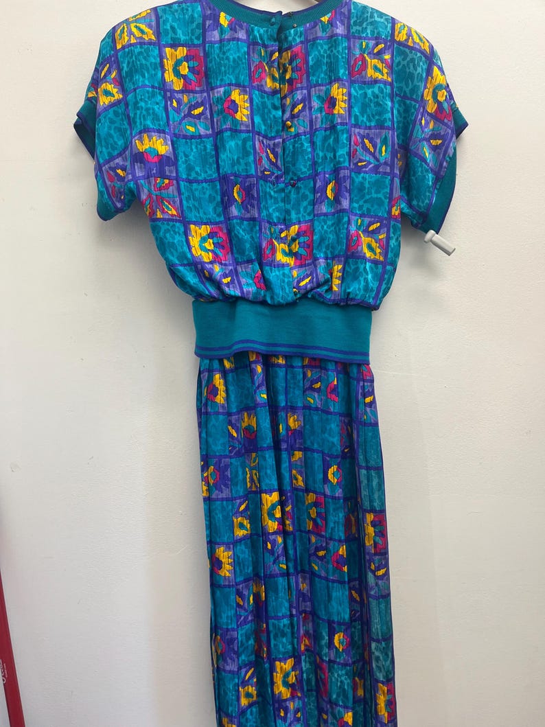 May include: A vintage teal dress with a square pattern in purple, yellow, and pink. The dress features short sleeves, a cinched waist, and a button-up back. The skirt is a long, flowing style.
