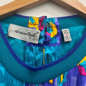 May include: Close-up of a vintage Adrianna Papell silk top. The top features a vibrant pattern in teal, purple, yellow, and pink. The neckline is ribbed teal and blue. The tag reads "100% Silk, Size 8, Made in Korea."