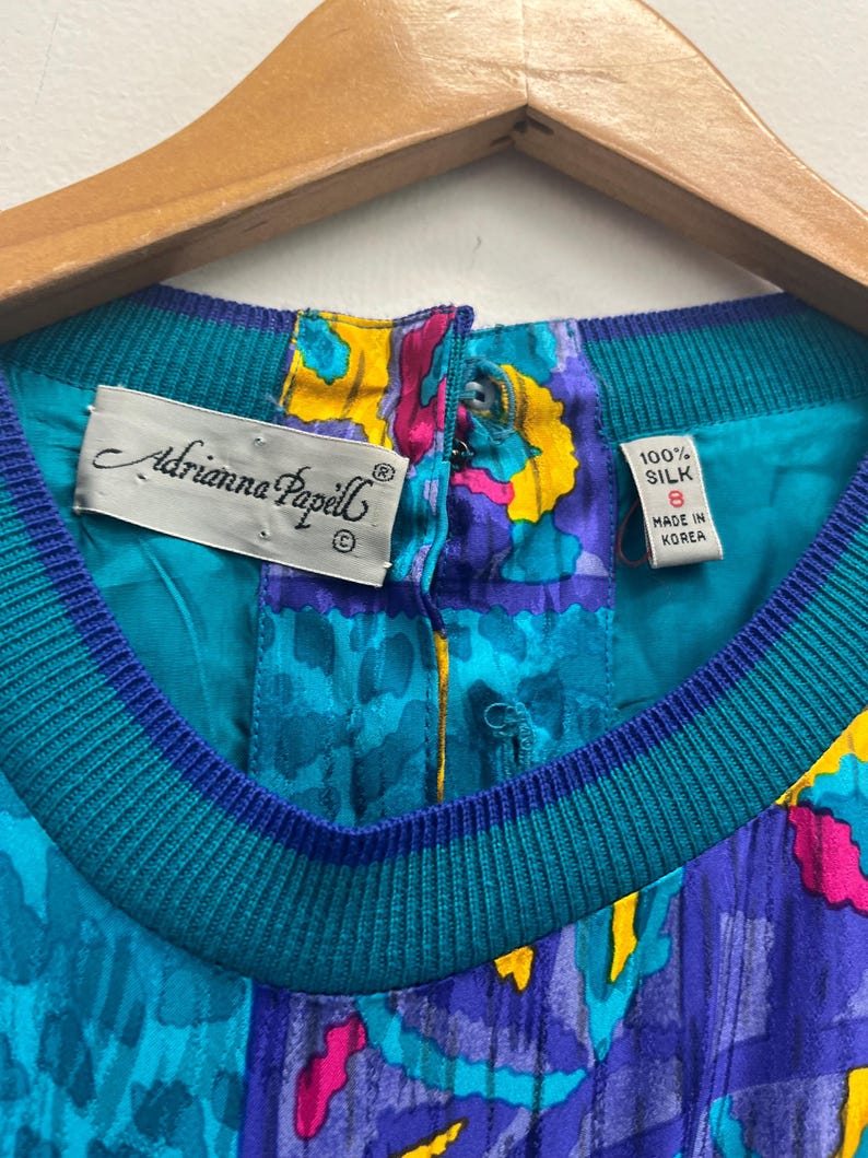 May include: Close-up of a vintage Adrianna Papell silk top. The top features a vibrant pattern in teal, purple, yellow, and pink. The neckline is ribbed teal and blue. The tag reads "100% Silk, Size 8, Made in Korea."