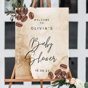 May include: A baby shower welcome sign on a wooden easel. The sign features a watercolour background with coffee beans and the text "Welcome to Olivia's Baby Shower 16.06.23."