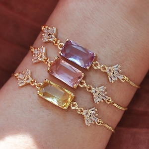 May include: Three gold-colored bracelets with rectangular, colorful gemstones. Each bracelet features a different gemstone color: yellow, pink, and purple. The bracelets are adorned with small, sparkling accents.