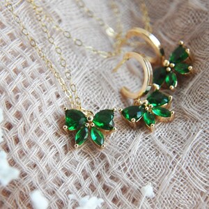 Puede incluir: Juego de joyas doradas que incluye un collar y pendientes. El collar tiene un colgante con detalles de piedras preciosas verdes en forma de mariposa. Los pendientes son de estilo aro con detalles de mariposa de piedras preciosas verdes a juego.