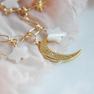 May include: A gold-colored charm bracelet featuring celestial-themed charms. The bracelet includes a crescent moon, stars, and a sun, all adorned with small crystals. The charms are suspended from a delicate chain.
