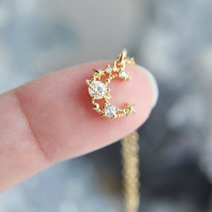 May include: A gold-coloured necklace with a crescent moon and star pendant. The pendant is decorated with clear, sparkling gemstones. The necklace is delicate and has a fine chain.