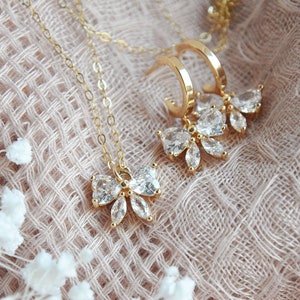 K&ouml;nnte beinhalten: Ein goldfarbenes Schmuckset mit Halskette und Ohrringen. Die Halskette hat eine feine Kette mit einem schleifenf&ouml;rmigen Anh&auml;nger, der mit klaren Kristallen verziert ist. Die Ohrringe sind im Creolen-Stil gehalten, ebenfalls mit schleifenf&ouml;rmigen Kristallanh&auml;ngern. Das Schmuckset wird auf einer strukturierten, hellrosa Oberfl&auml;che pr&auml;sentiert.