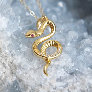 May include: A gold-colored snake pendant necklace. The snake has a red gem eye and is adorned with small, clear gems. The necklace is on a delicate gold chain. The background is a light blue, crystalline surface.