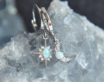 May include: Silver-toned earrings featuring a starburst and crescent moon design. Each earring has a central opal-like stone and is accented with small, clear crystals. The earrings are displayed against a light blue, crystalline background.