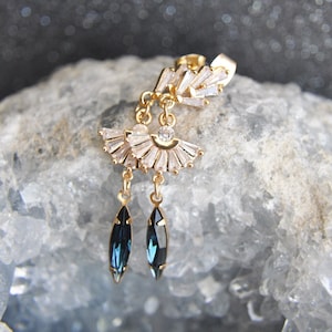 May include: Gold-toned earring with a fan-shaped design of clear crystals and two dangling, dark blue, teardrop-shaped crystals. The earring is set against a light blue, textured background.