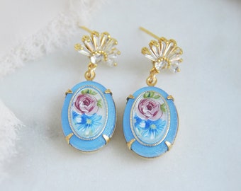May include: Gold-tone earrings with light blue oval pendants. Each pendant displays a hand-painted floral design with a pink rose and blue flowers. The earrings have a crystal and gold fan-shaped top.