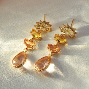 May include: A pair of gold-toned earrings with a sunburst design at the top. Each earring features yellow, orange, and peach-colored gemstone accents, culminating in a teardrop-shaped peach-colored stone.