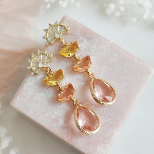 May include: Gold-toned drop earrings with a fan-shaped top, yellow and peach-colored accents, and teardrop-shaped pink gems. The earrings are displayed on a pink marble surface, showcasing their elegant design and color palette.