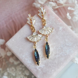 May include: Elegant gold-toned earrings with a fan-shaped design, adorned with clear crystals and a dangling dark blue teardrop-shaped stone. The earrings are displayed on a pink surface, showcasing their intricate details and sparkling appearance.