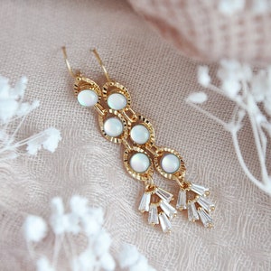 May include: Gold-toned dangle earrings featuring a series of circular opal-like stones connected by a chain design. The earrings have a cluster of baguette-cut crystals at the bottom. The background is a soft, neutral color.