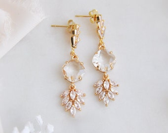 May include: A pair of gold-coloured drop earrings. Each earring features a large, round, clear crystal above a cluster of smaller, clear crystals in a leaf-like arrangement. The earrings have a post-back closure.