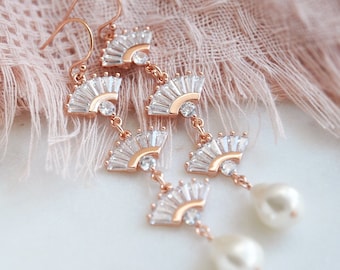 May include: Elegant rose gold earrings with a fan-shaped design, adorned with clear crystals and a dangling white pearl. The earrings are displayed against a soft pink fabric, showcasing their intricate details and delicate craftsmanship.