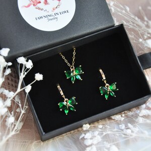 Puede incluir: Un conjunto de joyas en una caja negra, con un collar y pendientes dorados. Cada pieza tiene un dise&ntilde;o en forma de lazo con piedras preciosas de color verde esmeralda. La caja tiene una pegatina blanca con el texto "FAWNING IN LOVE Jewelry".