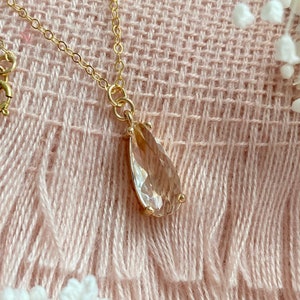 May include: A gold-colored necklace with a teardrop-shaped, light-brown gemstone pendant. The pendant is faceted and set in a gold bezel. The necklace is displayed on a pink, textured surface with white floral accents.
