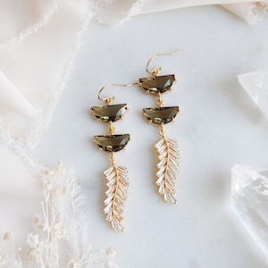 May include: Gold-tone drop earrings featuring three smoky quartz-colored, half-moon shaped stones. Below the stones are delicate, crystal-like, leaf-shaped accents. The earrings have a classic hook closure and are displayed on a white surface.