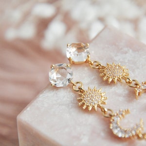 May include: Gold-toned earrings featuring clear, square-cut gemstones and celestial charms. The earrings have a sun and crescent moon design, with small, sparkling accents. The earrings are displayed on a pink marble surface.