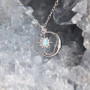 Puede incluir: Un collar de plata con un dise&ntilde;o celestial. El colgante incluye una luna creciente, una estrella con un &oacute;palo central y una peque&ntilde;a estrella. El collar se muestra sobre un fondo cristalino azul claro.