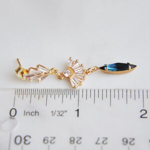 May include: A gold-toned earring with a fan-shaped design of clear crystals and a dangling blue and black crystal. The earring is approximately 2 inches long, as measured by the ruler in the image. The earring has a post back.